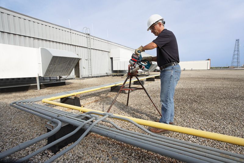 A person wearing safety equipment cuts a pipe with a cordless band saw on a rooftop.