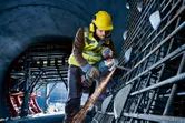 A worker wearing safety equipment grinds rebar with a cordless angle grinder in a tunnel.