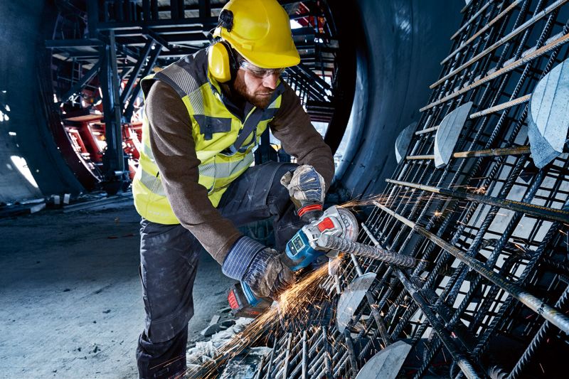 A person wearing safety equipment uses a cordless angle grinder to cut metal rebar.