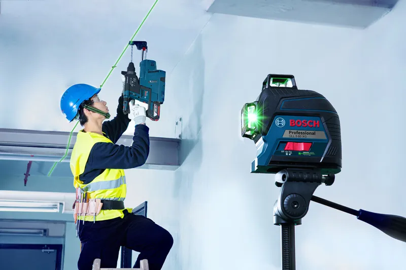 A person wearing safety equipment drills into a ceiling guided by a laser leveling tool.