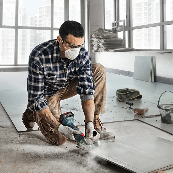 GWS 180-LI A person wearing safety equipment cuts a tile sheet with a power tool indoors.