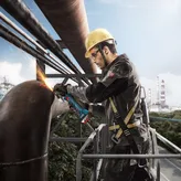 A person wearing safety equipment grinds a large metal pipe with an angle grinder.