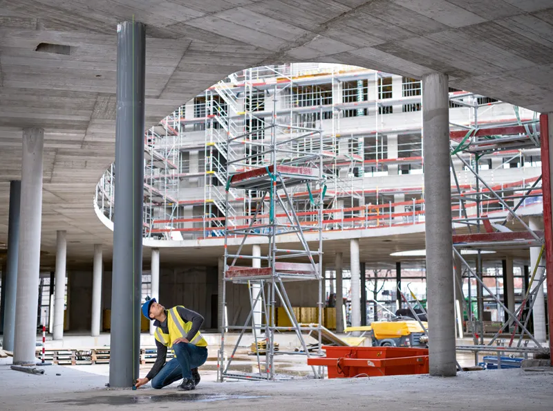 A person wearing safety equipment uses a laser measure on a column at a construction site.