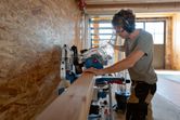 A person wearing safety equipment cuts a wooden beam using a cordless mitre saw.