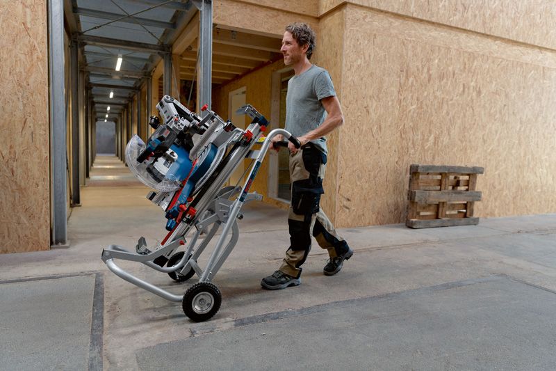 A person wearing safety equipment transports a cordless mitre saw on a wheeled stand.