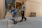 A person wearing safety equipment transports a cordless mitre saw on a wheeled stand.