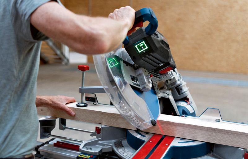 Person operates a cordless mitre saw to cut a wooden plank on a workshop table.