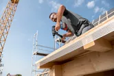 A person wearing safety equipment drills into a wooden beam on a building under construction.