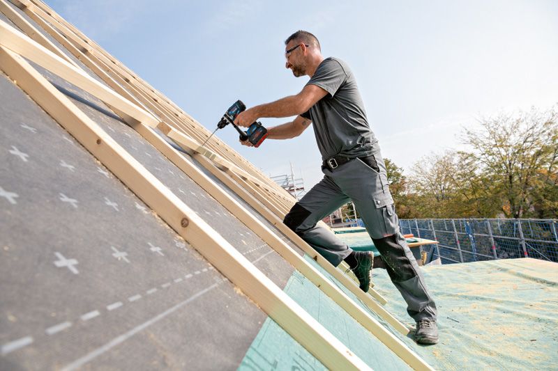 A person wearing safety equipment uses a cordless drill to fasten wood on a sloped roof.
