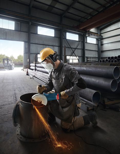 GWS 24-180 JZ A person wearing safety equipment uses an angle grinder to cut a large metal pipe indoors.