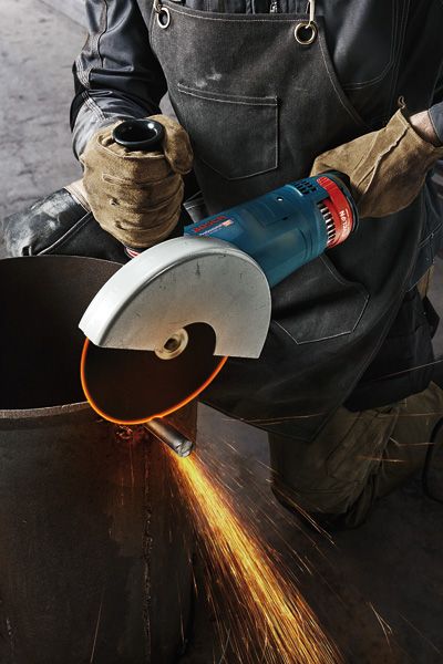 GWS 24-180 JZ A person wearing safety equipment cuts a metal pipe with a large angle grinder, sparks flying.