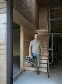 A person holding a mitre saw stands on wooden stairs at a construction site.