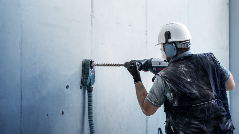Person wearing safety equipment drills into a concrete wall with a heavy-duty tool.