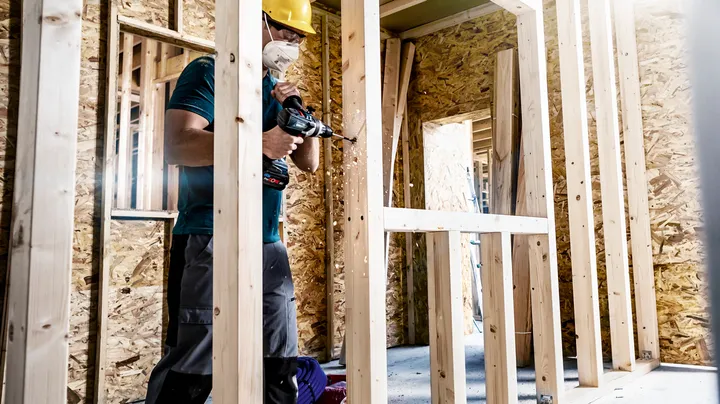 Object #200360549: 2793185_web_001 Person wearing safety equipment drills into a wooden frame at a construction site.