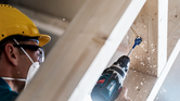 Worker wearing safety equipment drills a hole into a wooden beam.