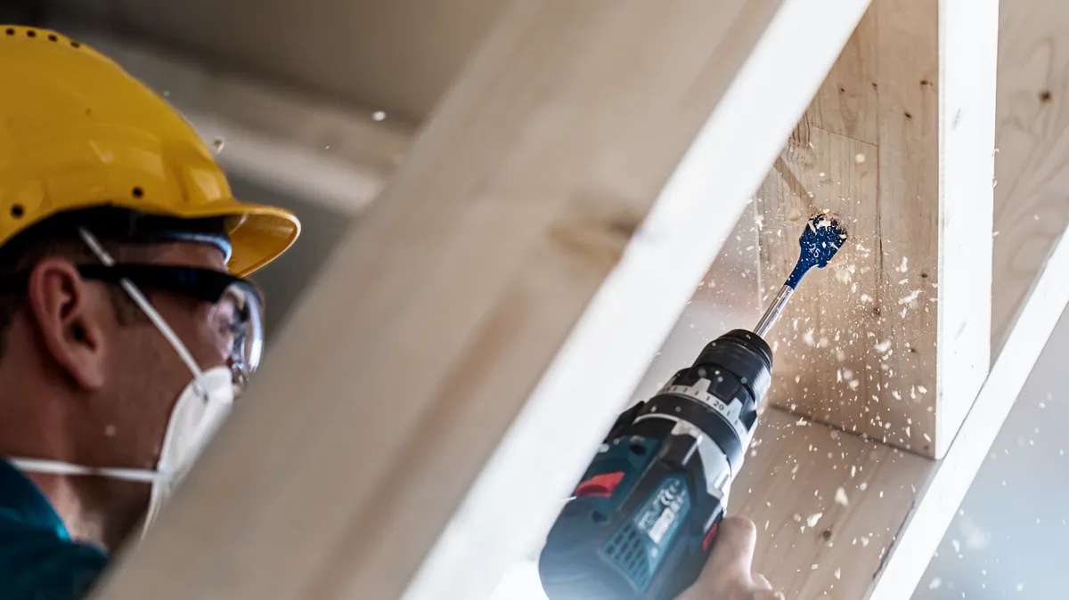 Worker wearing safety equipment drills a hole into a wooden beam.