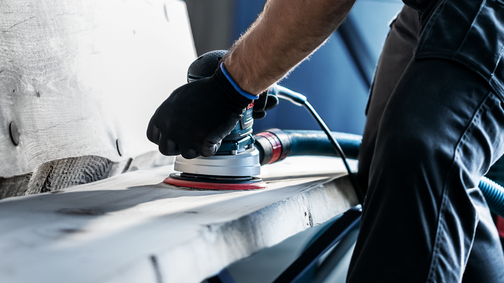 Person wearing safety equipment sanding a wooden surface with a power sander.