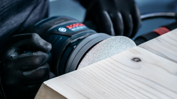 Worker wearing safety equipment sanding a wooden plank with a power sander.
