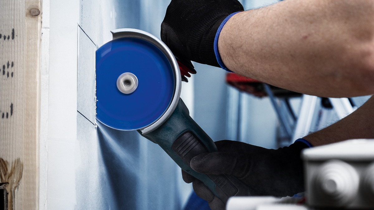 Worker wearing safety equipment cuts a square opening in drywall with a blue angle grinder.