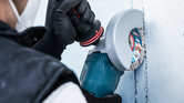 A worker wearing safety equipment uses an angle grinder to cut a channel in a wall.