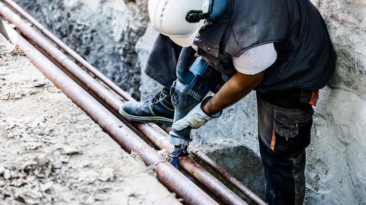 Object #200360737: 2793318_web_001 Person wearing safety equipment drills into a pipe in an outdoor trench.