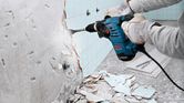A person wearing safety equipment uses a rotary hammer to remove tiles from a wall.