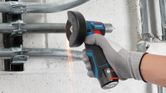A person wearing safety equipment uses a cordless angle grinder to cut metal pipes.