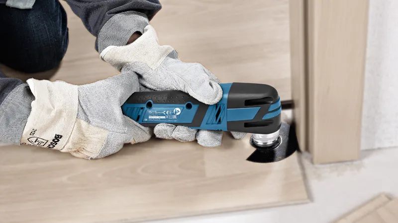 A person wearing safety equipment trims a wooden floorboard with a cordless multi-cutter.