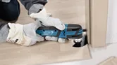 A person wearing safety equipment trims a wooden floorboard with a cordless multi-cutter.