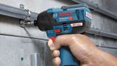 A person fastens a metal pipe to a wall using a cordless impact driver.