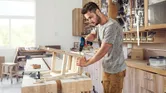 A person assembles a wooden chair using a cordless drill in a workshop.
