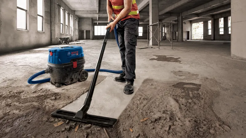GAS 12-25 A person wearing safety equipment vacuums concrete dust in a large unfinished building.