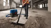 A person wearing safety equipment vacuums concrete dust in a large unfinished building.