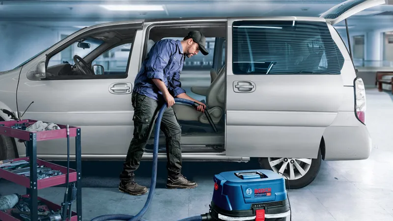 GAS 12-25 A person cleans a car interior with a vacuum next to a tool cart in a garage.