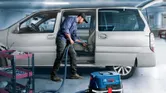 A person cleans a car interior with a vacuum next to a tool cart in a garage.