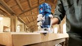 A person wearing safety equipment uses a palm router to shape the edge of a wooden board.