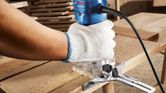 A person wearing safety equipment trims wood with a palm router on a workbench.