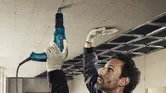 A person wearing safety equipment secures drywall to a ceiling using a screwdriver.