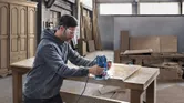 A person wearing safety equipment uses a router on a wooden tabletop in a workshop.