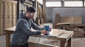 A person wearing safety equipment uses a router on a wooden tabletop in a workshop.