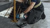 A person wearing safety equipment tightens a bolt on a metal post using a cordless impact wrench.
