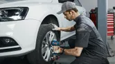 A person wearing safety equipment uses a cordless impact wrench to tighten wheel bolts on a car.