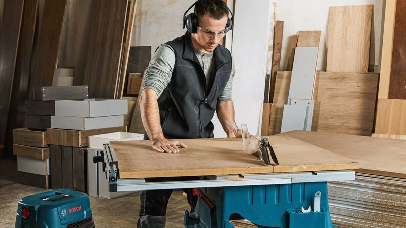 A person wearing safety equipment cuts wood using a table saw in a workshop.