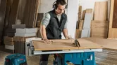 A person wearing safety equipment cuts wood using a table saw in a workshop.
