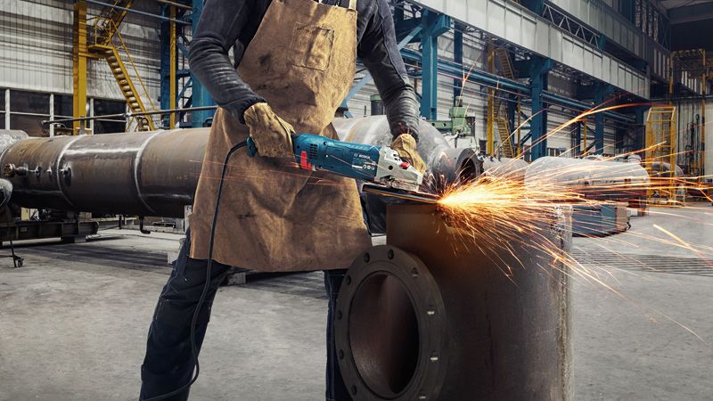 GWS 24-180 JZ A person wearing safety equipment grinds a metal pipe with an angle grinder, sparks flying.
