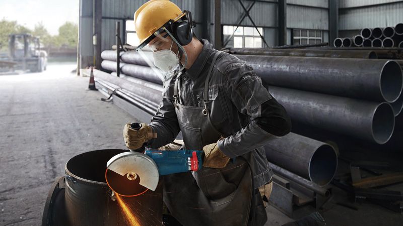 GWS 24-180 JZ A worker wearing safety equipment grinds a metal pipe with a large angle grinder.