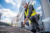 A person wearing safety equipment uses a cordless drill driver on construction ground.