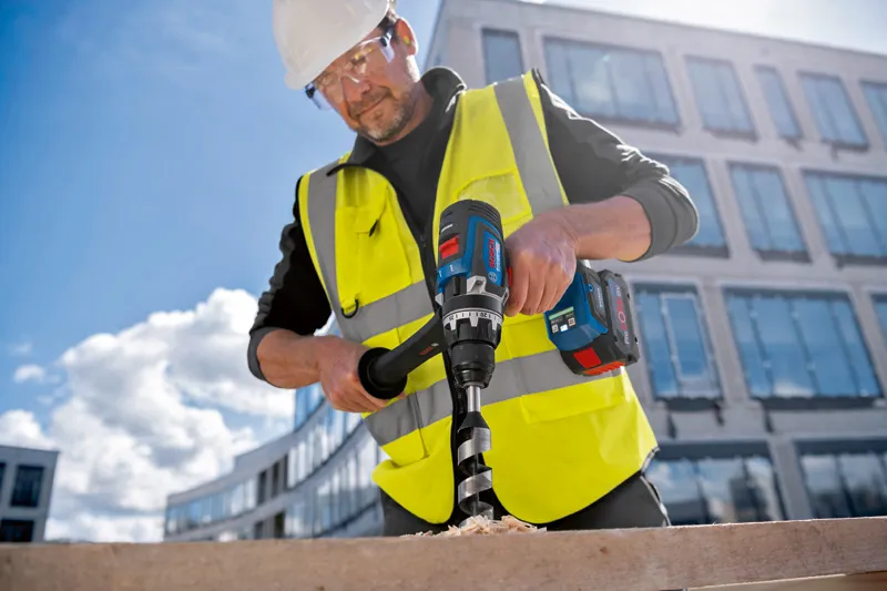 A person wearing safety equipment drills into wood with a cordless drill at a construction site.