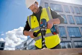 A person wearing safety equipment drills into wood with a cordless drill at a construction site.