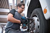 A person wearing safety equipment uses a cordless impact wrench to tighten truck wheel bolts.